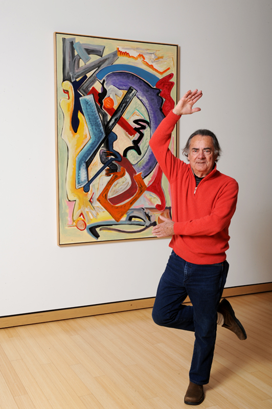 Bill Barrett posing on one leg with one arm in the air as if he were dancing and next to his artwork Oil Painting Number 2 which has colorful abstract musical symbols