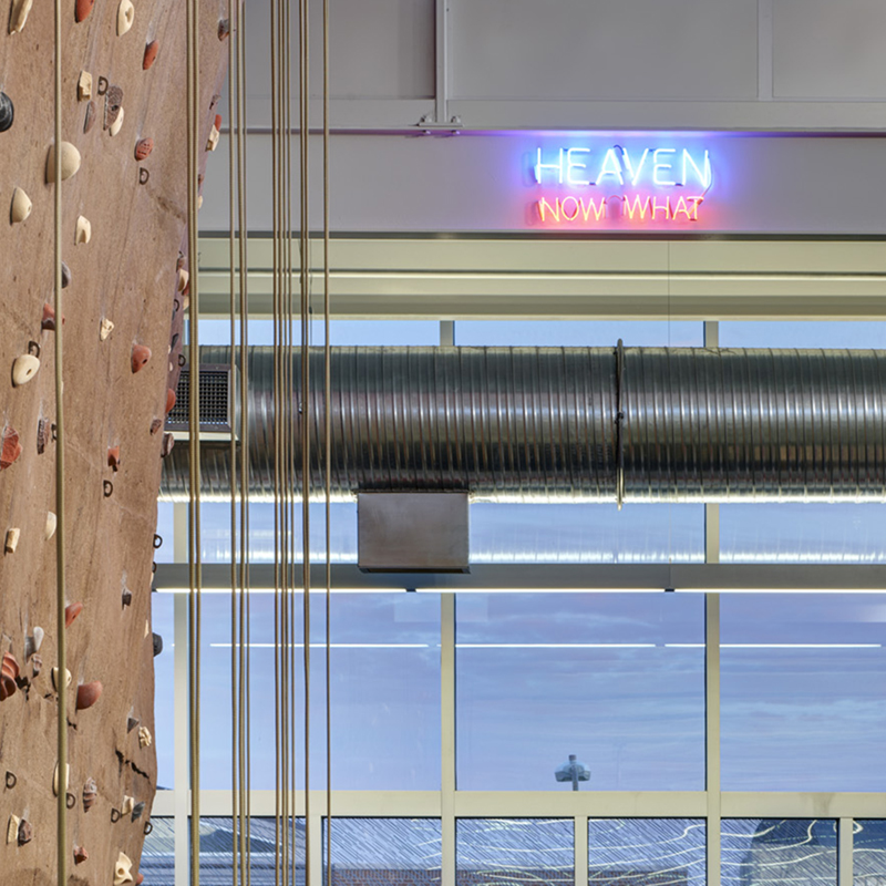 The rock-climbing wall at State Gymnasium with a neon sign in the background saying Heaven Now What