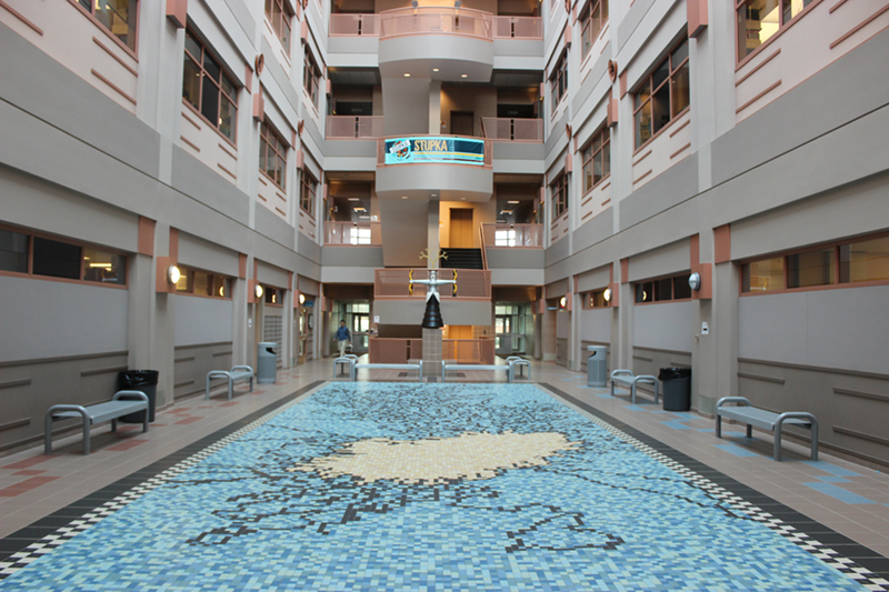 Photo of the floor of the Molecular Biology Building taken from the ground level. The mosaic floor tiles look like a swimming pool. There is a metal sculpture at the end holding things in its hands.