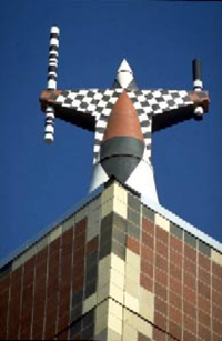 Closeup of the sculpture on the roof corner of the Molecular Biology Building. A figure is covered in black and white squares with some maroon accent colors. It is holding two black and white rods