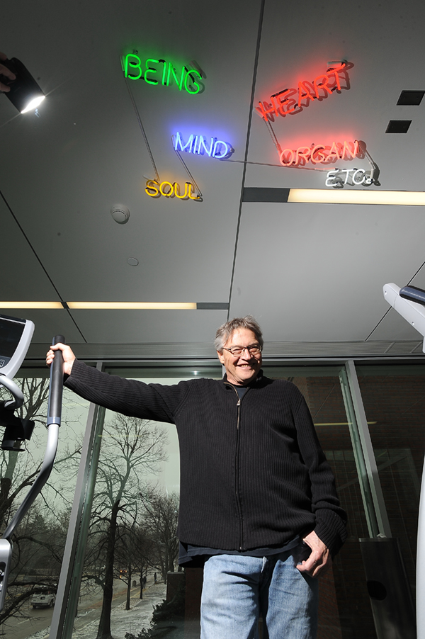Terry Allen with his hand on an elliptical machine handle and neon words on the ceiling reading Being, Mind, Soul, Heart, Organ, Etc.