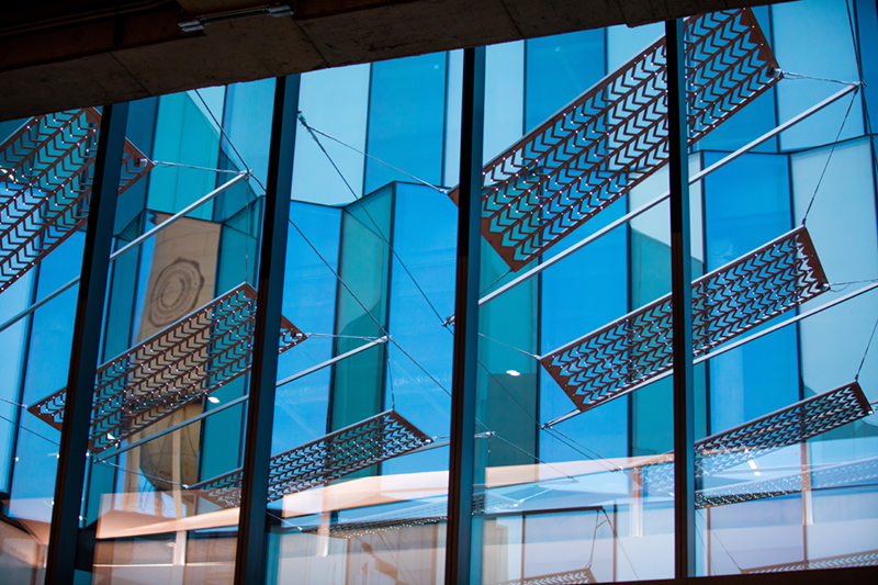 Photo taken from inside looking up and out windows into a courtyard with suspended copper panels and large glass pleated windows in the background