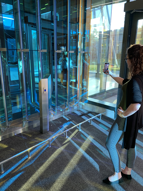 Photo of a person with a phone taking a selfie in front of pleated dichroic glass reflecting greens and blues