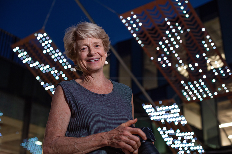 Photo of Catherine Widgery smiling and holding a camera in front of panels with twinkling lights